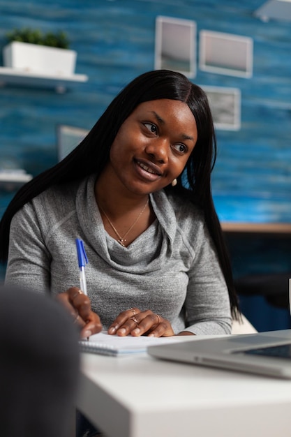 Afro-Amerikaanse vrouw schrijft informatie over leerboekbestand met pen, kijkend naar laptop. Persoon die aantekeningen maakt op notebookpapier en op afstand werkt. Volwassen ontwerpen van zakelijk project.