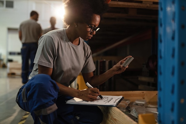 Afro-Amerikaanse vrouwelijke werknemer die producten controleert vóór de verzending en aantekeningen maakt in een magazijn