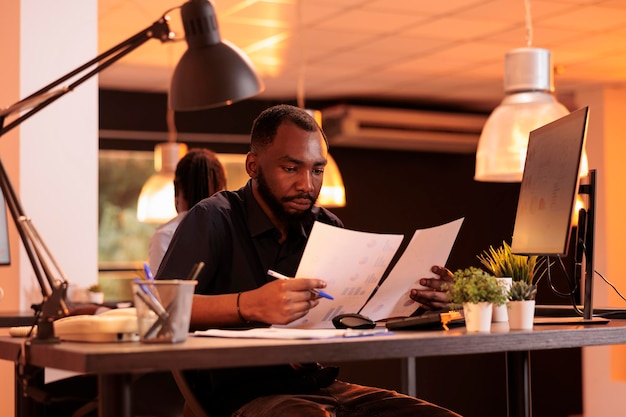 Afro-Amerikaanse werknemer maakt aantekeningen over contractrapport, analyseert papierwerk met zakelijke gegevens op kantoor. Werken met documenten om onderzoek te doen en uitvoerende oplossingen te vinden.