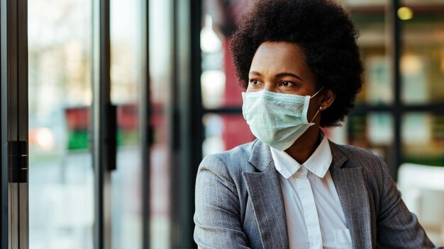 Afro-Amerikaanse zakenvrouw die een beschermend masker op haar gezicht draagt terwijl ze in een café zit en door het raam kijkt