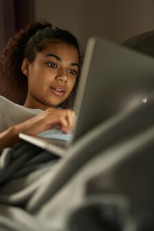 Afstand leren. aantrekkelijke jonge vrouwelijke student van gemengd ras die studeert met haar laptop terwijl ze thuis op bed ligt