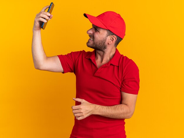 Agressieve jonge blanke bezorger in rood uniform en pet verhogen en kijken naar mobiele telefoon geïsoleerd op oranje muur