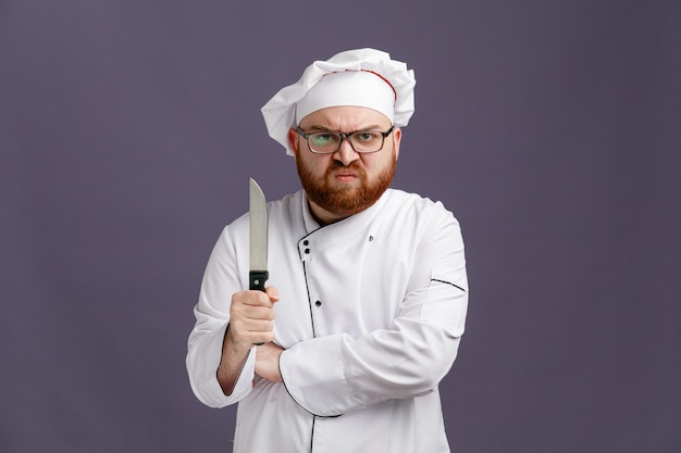 Agressieve jonge chef-kok met een uniforme bril en een pet die een mes vasthoudt terwijl hij naar de camera kijkt terwijl hij de hand onder de arm houdt geïsoleerd op een paarse achtergrond