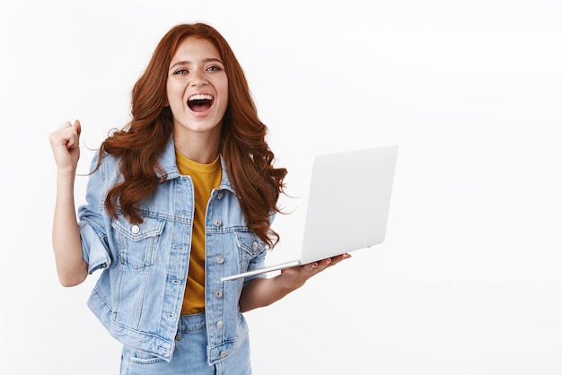 Ambitieus roodharig meisje in spijkerjasje houden laptop gelukkig glimlachen bereiken opgelucht prestatie zegevieren van goed nieuws winnen loterij vrolijk winnen witte achtergrond