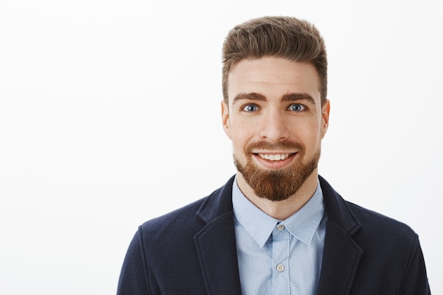 Ambitieuze knappe en jonge stijlvolle volwassen man met baard en grote blauwe ogen glimlachend opgewonden en tevreden grijns staande in trendy pak over grijze muur wachten op kans vaardigheden tonen over grijze muur