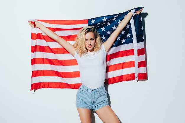 Amerikaanse blonde vrouw die de vlag van de VS houdt die over een witte muur wordt geïsoleerd