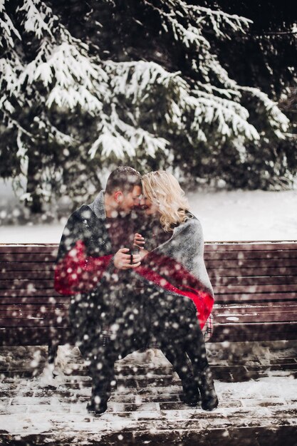 Amoureuze paar vriendje en vriendin poseren en zittend op een bankje in het winterbos. Mooie blonde en glimlachende man die naar de camera kijkt en kopjes met koffie in handen houdt. Begrip liefde.