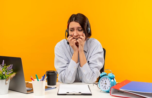 Angstig jong call centreermeisje die hoofdtelefoonszitting bij bureau dragen die laptop bekijken en haar vingers bijten die op oranje worden geïsoleerd