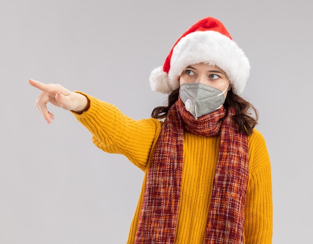 Angstig jong Slavisch meisje met kerstmuts en met sjaal om de nek die medisch masker draagt ?? kijkend en wijzend naar kant geïsoleerd op een witte achtergrond met kopie ruimte