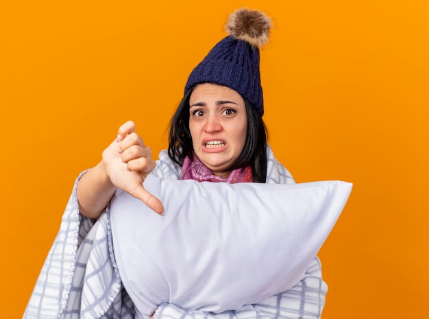 Gratis foto angstig kaukasisch ziek meisje dragen winter hoed en sjaal gewikkeld in geruite kussen houden kijken camera weergegeven: duim omlaag geïsoleerd op een oranje achtergrond