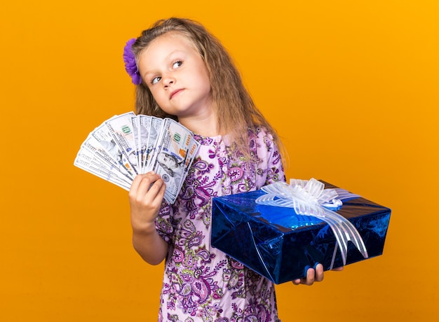 angstig klein blond meisje met geschenkdoos en geld kijkend naar kant geïsoleerd op oranje muur met kopieerruimte