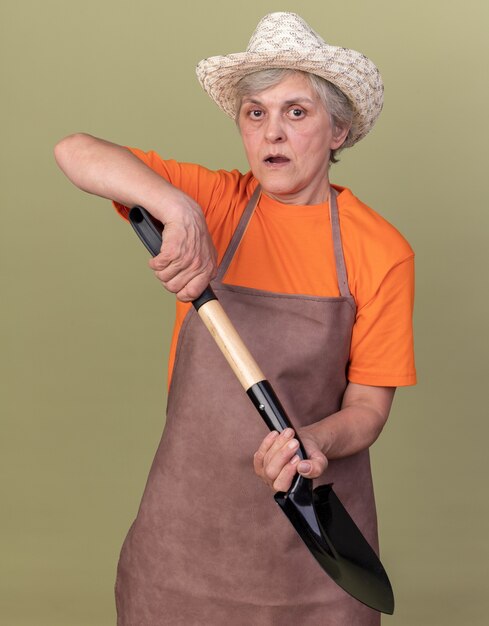 Angstige bejaarde vrouwelijke tuinman die het tuinieren de schop van de hoedholding op olijfgroen draagt