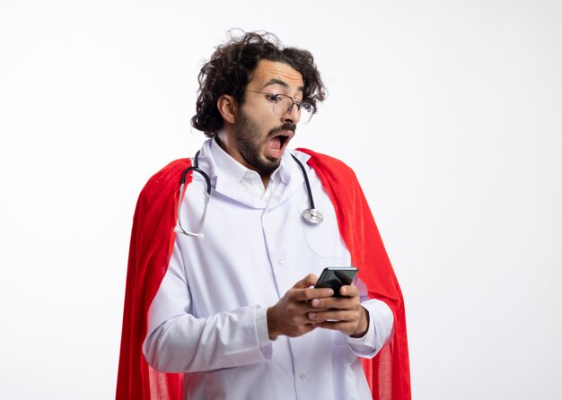 Angstige jonge blanke superheld man in optische bril met doktersuniform met rode mantel en met stethoscoop om nek houdt vast en kijkt naar telefoon