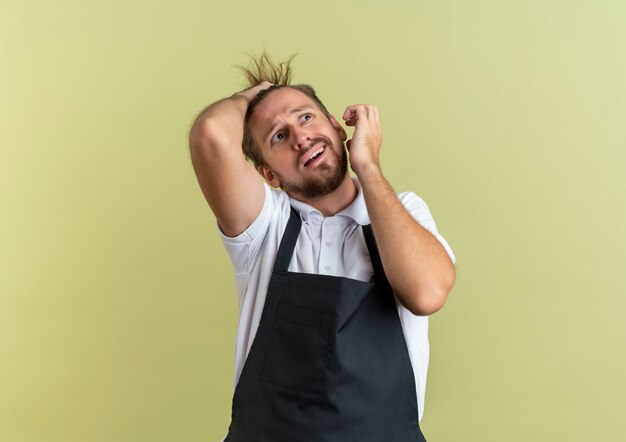 Angstige jonge knappe kapper die kant kijkt die zijn haar aanraakt met een andere hand die in de lucht wordt gehouden geïsoleerd op olijfgroen met kopie ruimte