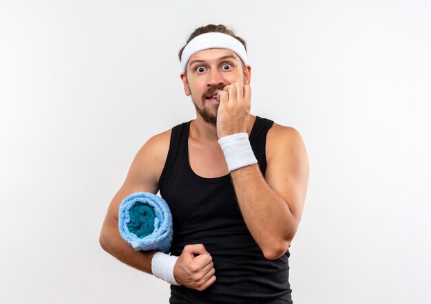 Angstige jonge knappe sportieve man met hoofdband en polsbandjes die een handdoek vasthouden en zijn vingers bijten geïsoleerd op een witte muur met kopieerruimte