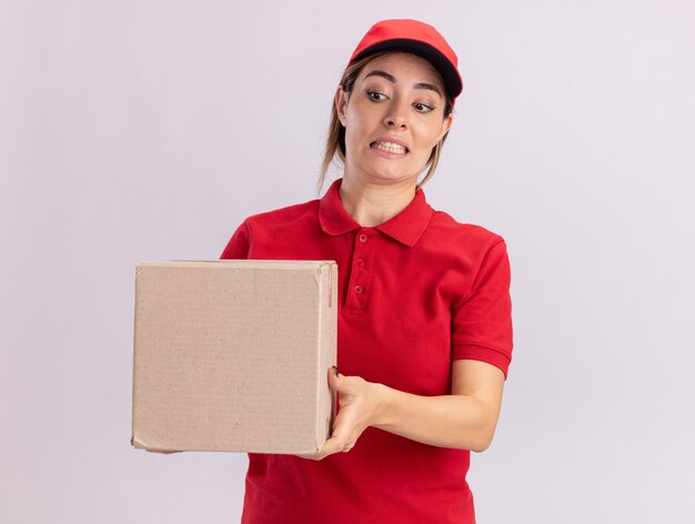 Angstige jonge mooie leveringsvrouw in uniform houdt en kijkt naar kartonnen doos geïsoleerd op een witte muur