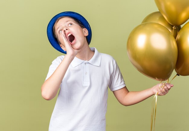 angstige jonge Slavische jongen met blauwe feestmuts die heliumballonnen vasthoudt en de hand dicht bij de mond houdt en iemand belt die omhoog kijkt geïsoleerd op olijfgroene muur met kopieerruimte