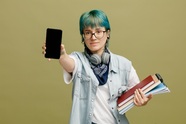 Angstige jonge vrouwelijke student met een bril, bandana en koptelefoon om de nek met notitieblokken die de mobiele telefoon uitrekken naar de camera die naar de camera kijkt die op een olijfgroene achtergrond wordt geïsoleerd