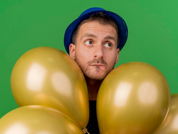 Angstige knappe man met blauwe feestmuts houdt helium ballonnen geïsoleerd op groene muur met kopie ruimte