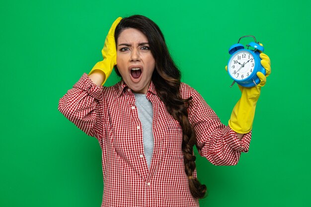 Angstige, mooie blanke schonere vrouw met rubberen handschoenen die haar hand op haar hoofd legt en een wekker vasthoudt