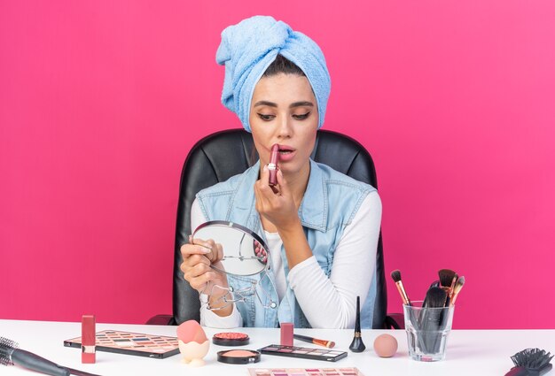 Angstige, mooie blanke vrouw met gewikkeld haar in een handdoek zittend aan tafel met make-uptools die naar een spiegel kijkt die lippenstift aanbrengt