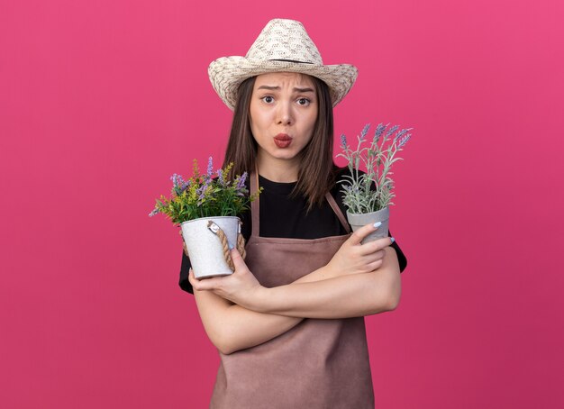 Angstige, mooie blanke vrouwelijke tuinman die tuinhoed draagt, staat met gekruiste armen met bloempotten geïsoleerd op een roze muur met kopieerruimte