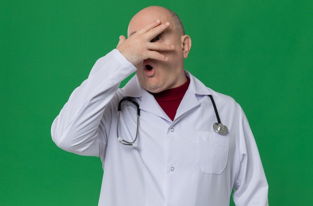 Angstige volwassen man in doktersuniform met stethoscoop die hand op zijn gezicht legt en door vingers kijkt