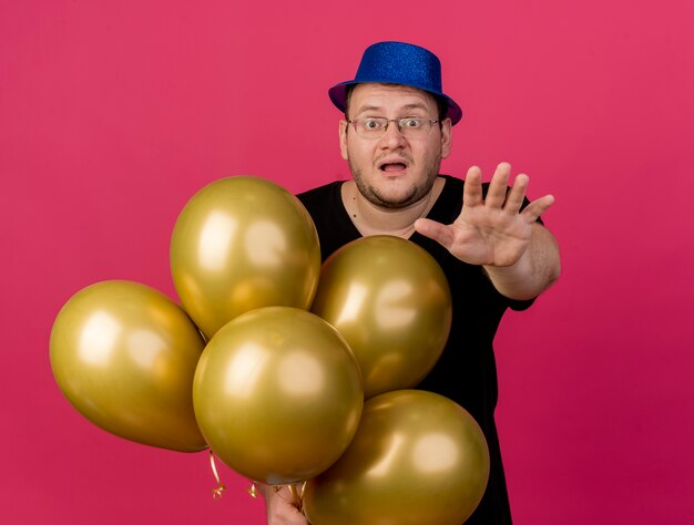 Angstige volwassen Slavische man met een optische bril met een blauwe feestmuts houdt heliumballonnen vast die hun hand uitstrekken