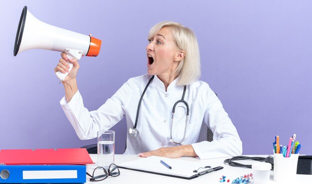 Angstige volwassen Slavische vrouwelijke arts in medische gewaad met stethoscoop zittend aan een bureau met kantoorhulpmiddelen schreeuwen in luidspreker kijkend naar kant geïsoleerd op paarse achtergrond met kopie ruimte