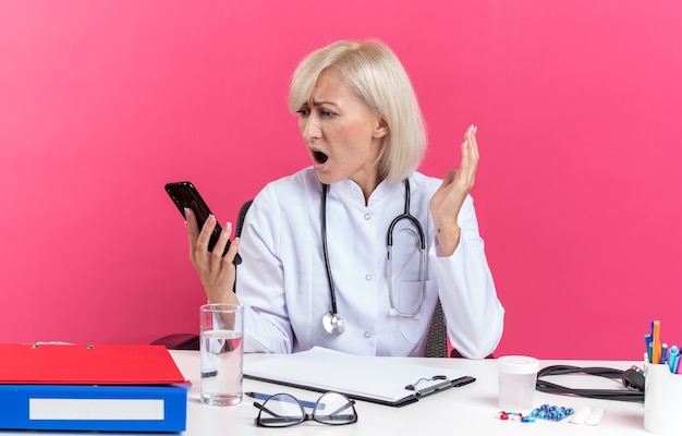 Angstige volwassen Slavische vrouwelijke arts in medische gewaad met stethoscoop zittend aan een bureau met office tools houden en kijken naar telefoon geïsoleerd op roze achtergrond met kopie ruimte