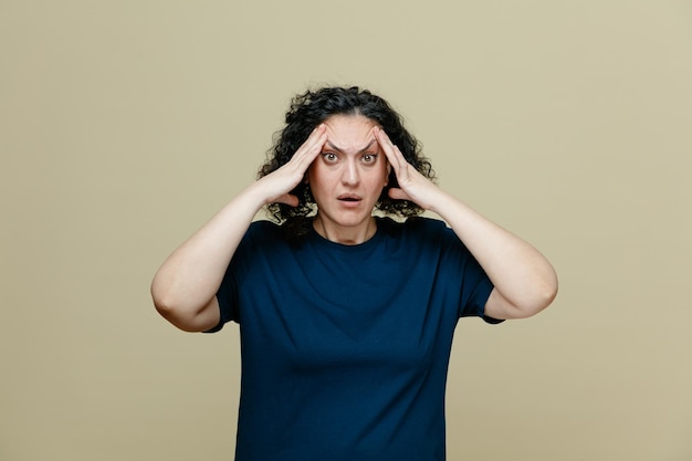 Angstige vrouw van middelbare leeftijd met een t-shirt die de handen op het hoofd houdt en naar de camera kijkt die op een olijfgroene achtergrond wordt geïsoleerd