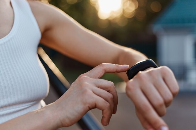 Anonieme vrouw die een draagbaar apparaat voor fitness en gezondheid controleert op haar hand, onbekende vrouw in witte top poseren buiten in het stadion bij zonsondergang.