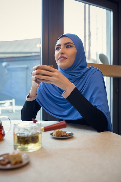 Apparaten gebruiken. Mooie Arabische vrouw bijeen in café of restaurant, vrienden of zakelijke bijeenkomst. Samen tijd doorbrengen, praten, lachen. Moslim levensstijl. Stijlvolle en vrolijke modellen met make-up.