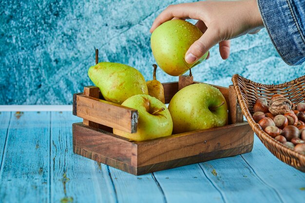 Appels en peer in houten mand en kom met hazelnoten op blauwe oppervlakte.