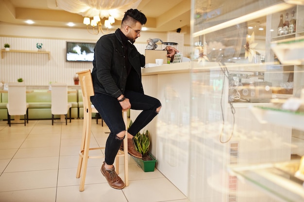 Arabische man draagt een zwarte spijkerjas en een bril in café drinkt koffie aan de bar met boek Stijlvolle en modieuze Arabische modelman