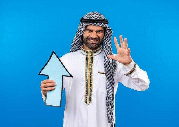 Arabische man in traditionele kleding die grote blauwe pijl met geïrriteerde uitdrukking op gezicht houdt die zich over blauwe muur bevinden