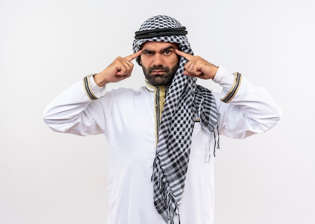 Arabische man in traditionele kleding die zijn slapen richt met een ernstige zelfverzekerde uitdrukking die over een witte muur staat