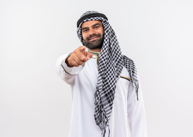 Gratis foto arabische man in traditionele kleding lachend met blij gezicht wijzend met vinger staande over witte muur