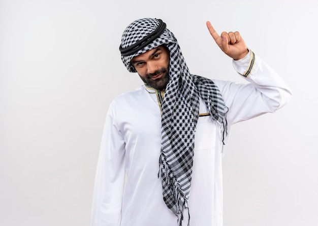 Arabische man in traditionele kleding met zelfverzekerde glimlach die met wijsvinger omhoog wijst die zich over witte muur bevindt