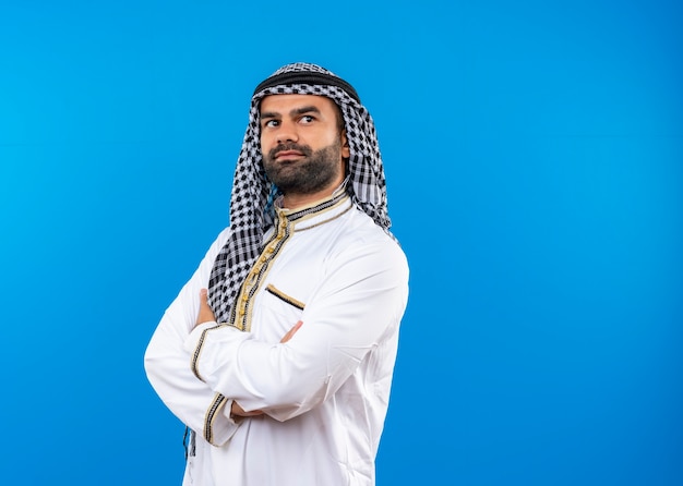 Gratis foto arabische man in traditionele kleding opzij kijken met zelfverzekerde uitdrukking met gekruiste handen op de borst staande over blauwe muur