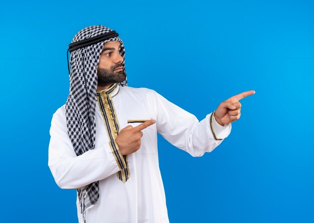 Arabische man in traditionele kleding opzij kijkend wijzend met de vingers opzij met een ernstig gezicht dat over de blauwe muur staat