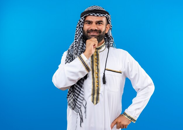 Arabische man in traditionele slijtage met glimlach op gezicht positief en blij staande over blauwe muur