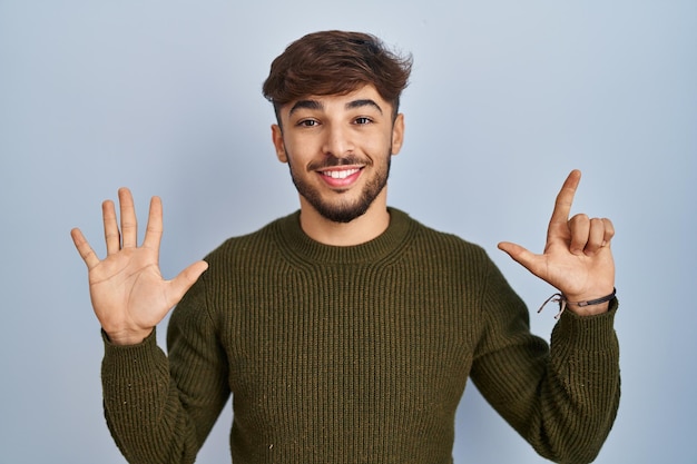 Arabische man met baard die over blauwe achtergrond staat en omhoog wijst met vingers nummer zeven terwijl hij zelfverzekerd en gelukkig glimlacht.