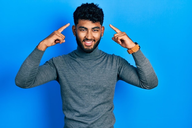 Gratis foto arabische man met baard met coltrui glimlachend wijzend naar het hoofd met beide handen vinger, geweldig idee of gedachte, goed geheugen