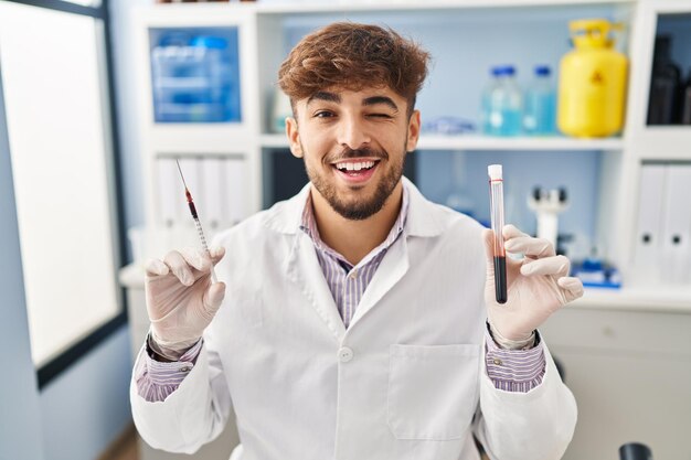 Arabische man met baard werken bij wetenschapper laboratorium houden bloedmonster knipogend kijken naar de camera met sexy uitdrukking vrolijk en blij gezicht
