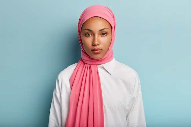 Arabische vrouw met kalme, ernstige uitdrukking, heeft een donkere huid, gehuld in roze sluier, draagt een wit overhemd, heeft geen make-up, natuurlijke schoonheid, modellen over blauwe muur. close-up shot van moslim dame