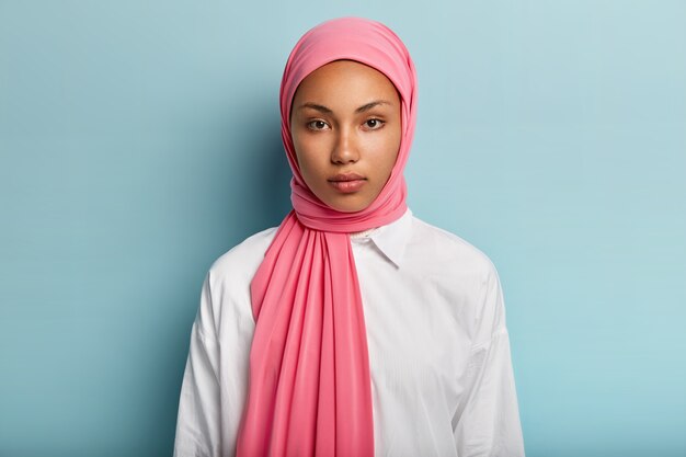 Arabische vrouw met kalme, ernstige uitdrukking, heeft een donkere huid, gehuld in roze sluier, draagt een wit overhemd, heeft geen make-up, natuurlijke schoonheid, modellen over blauwe muur. Close-up shot van moslim dame