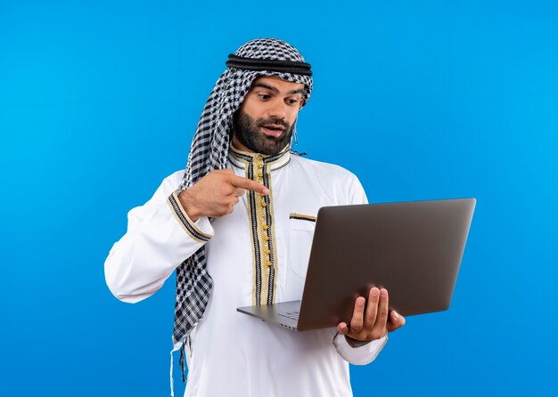 Arabische zakenman die in traditionele slijtage laptop houdt die ernaar kijkt met glimlach op gezicht die met vinger ernaar richt die zich over blauwe muur bevindt