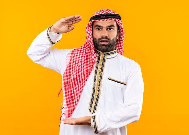 Arabische zakenman in traditionele kleding die verbaasd kijkt en groot gebaar toont, meetsymbool dat zich over oranje muur bevindt