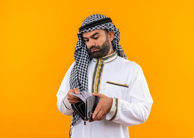 Arabische zakenman in traditionele slijtage die contant geld in zijn portefeuille bekijkt met ernstig gezicht dat zich over oranje muur bevindt